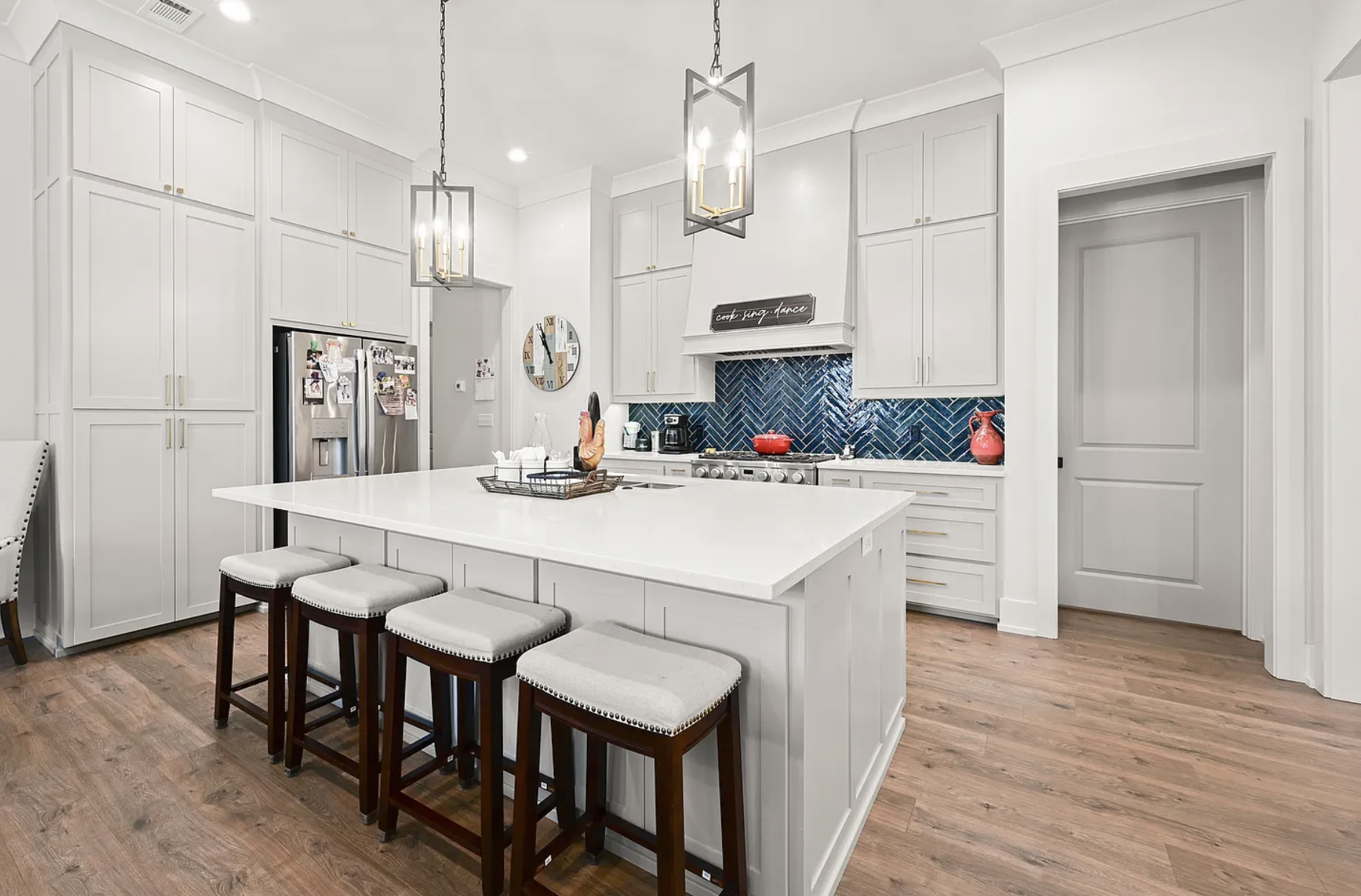 Luxury kitchen with white cabinets, white quartz countertops, stainless steel appliances, island sink, built-in stainless steel refrigerator, large range hood, and blue topaz herringbone diagonal backsplash tiles.