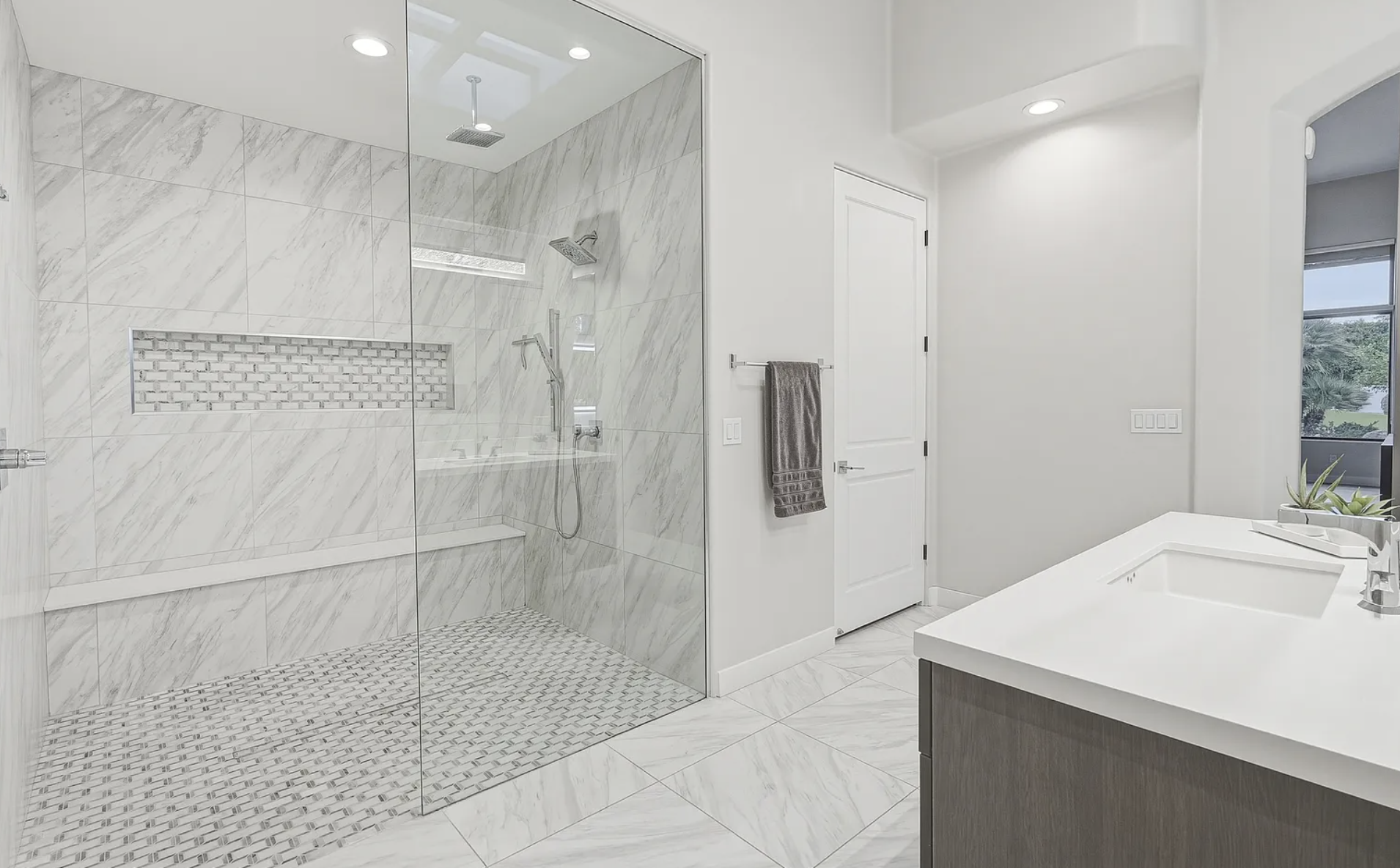 Luxury bathroom with a large glass shower enclosure, silver mosaic metal shower floor, overhead rain shower head, wall-mounted shower fixture, body sprays, recessed mosaic feature wall, and white vanity sink.