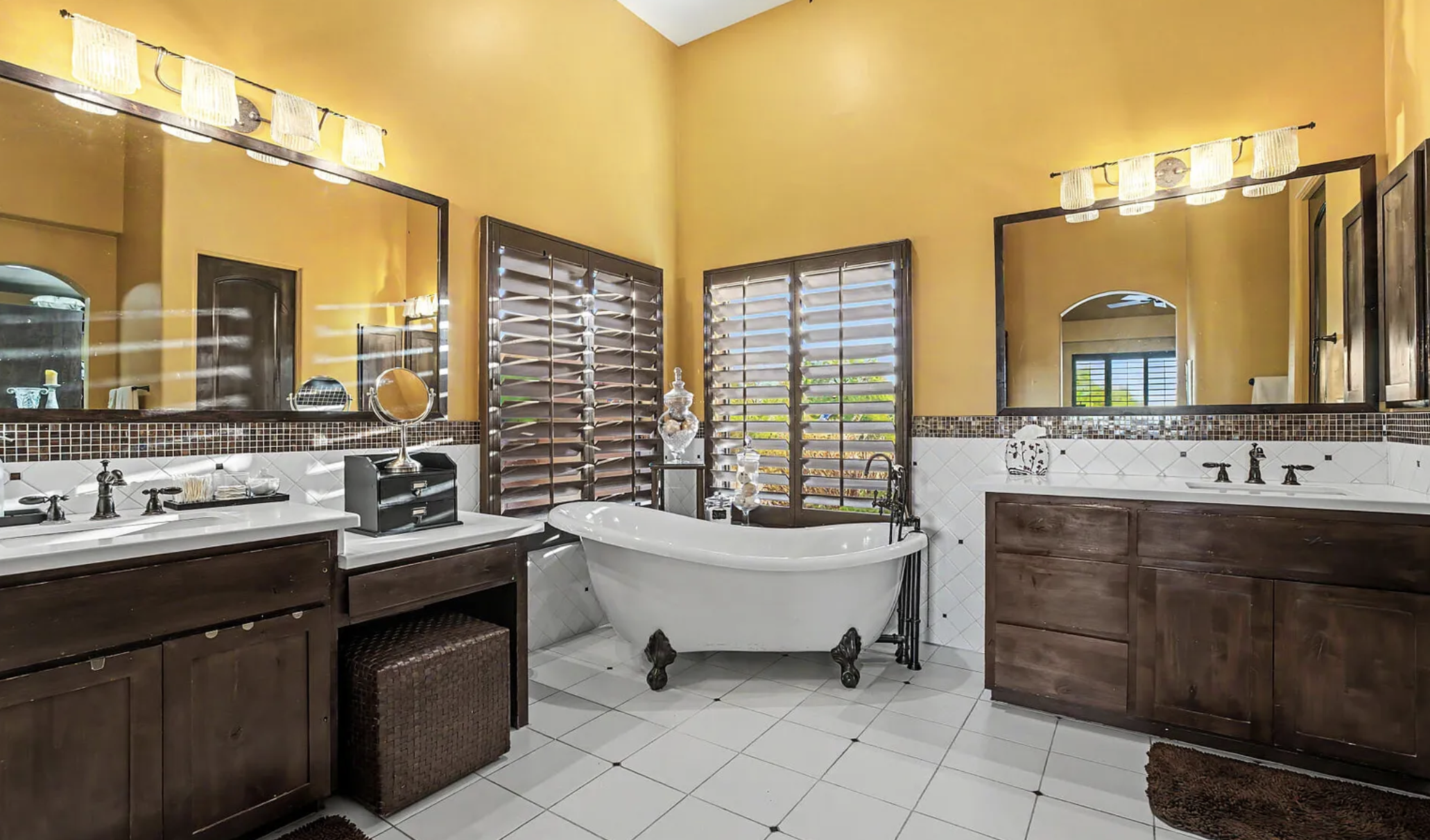 Luxury bathroom with white clawfoot tub and black feet, dark wood vanities, white quartz counters, oil rubbed bronze fixtures, dual sinks, mirrors, and windows with matching wood slats.