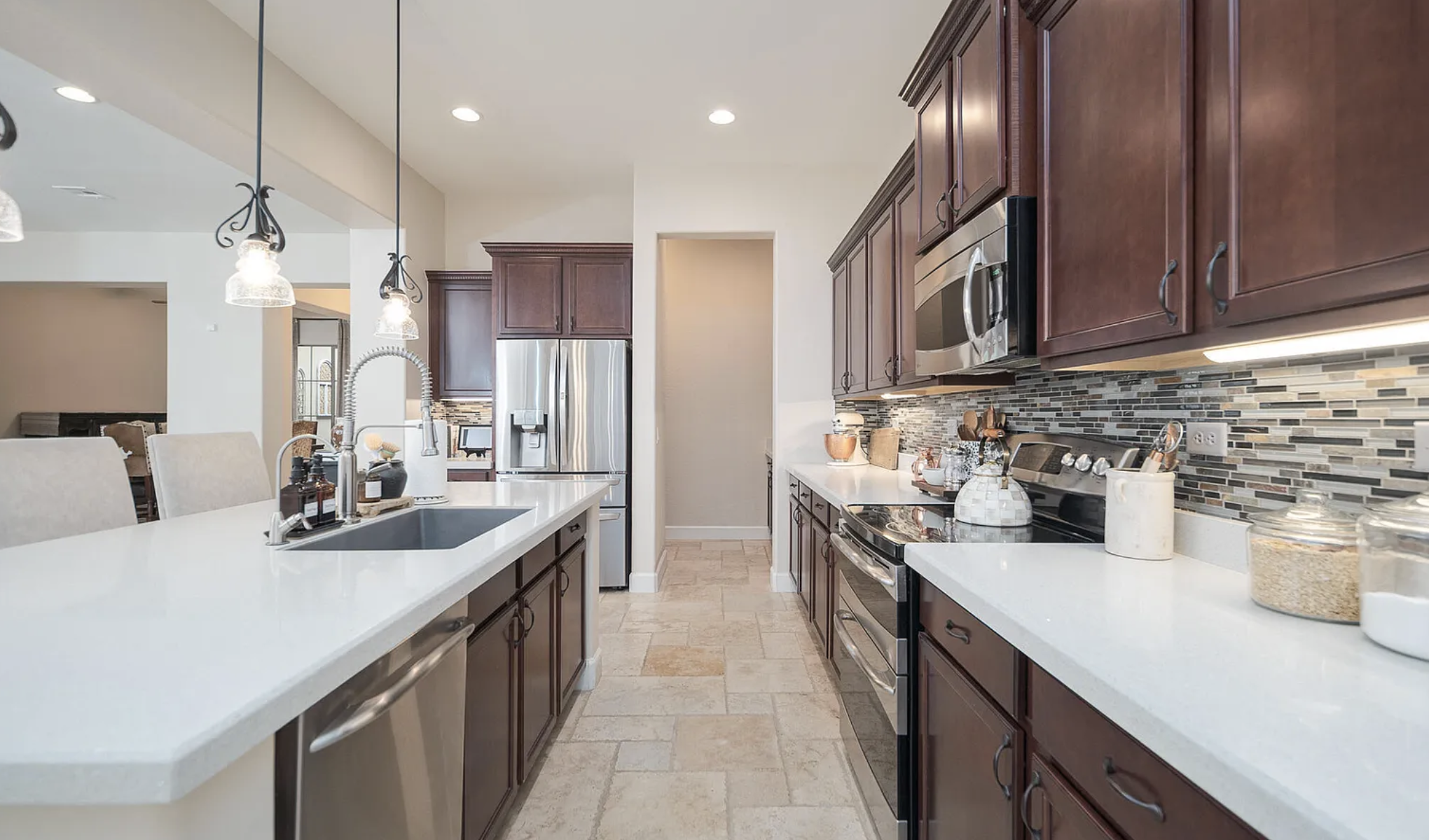 Modern kitchen with dark wood cabinets, stainless steel appliances, white quartz countertops, a large island with integrated sink and dishwasher, and grey black and white glass mosaic backsplash.